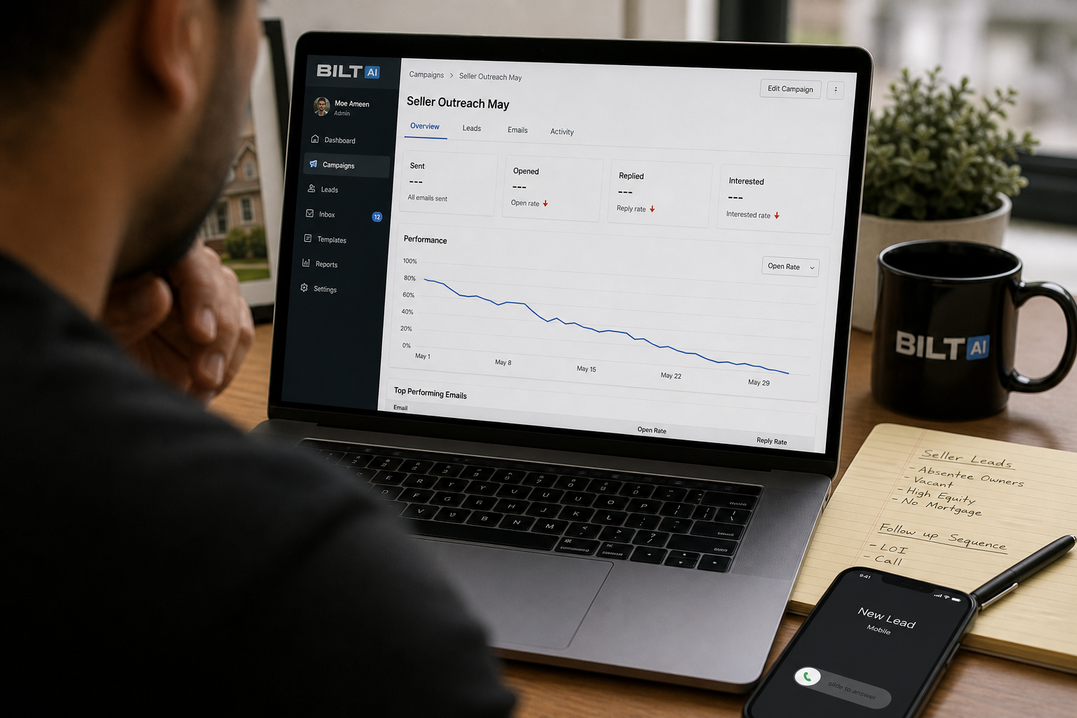over-the-shoulder view of a laptop showing an email campaign dashboard with declining open rates, real estate notes and a phone nearby, natural desk lighting